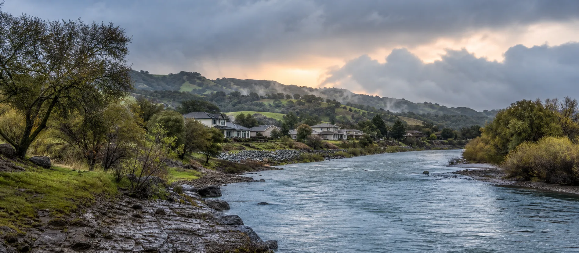 A full California river beneath atmospheric-river clouds near foothill homes.